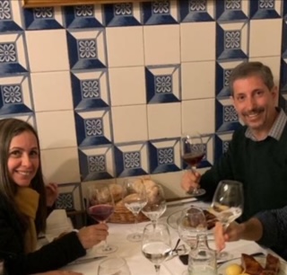 Casal sorridente brindando com taças de vinho em uma mesa do restaurante Sobrino de Botín em Madri, com parede de azulejos decorados ao fundo.