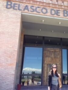 Mulher sorridente com óculos escuros posando em frente à entrada principal de tijolos ecológicos da vinícola Belasco de Baquedano.