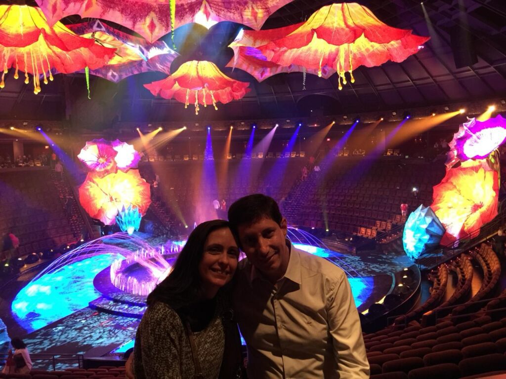 Casal sorrindo em primeiro plano dentro do teatro do show "O" do Cirque du Soleil em Las Vegas, com o palco aquático e decorações de flores gigantes iluminadas ao fundo.