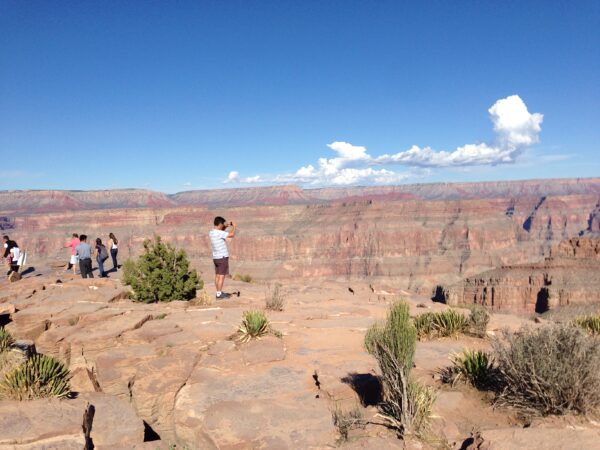 Turistas em um mirante rochoso observando a imensidão geológica e os paredões avermelhados do Grand Canyon, nos Estados Unidos, sob céu azul com nuvens.