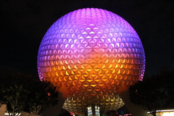 A grande esfera geodésica Spaceship Earth iluminada à noite em tons de roxo, rosa e laranja no parque Epcot Center em Orlando, Estados Unidos, com silhuetas de árvores na base contra o fundo escuro.