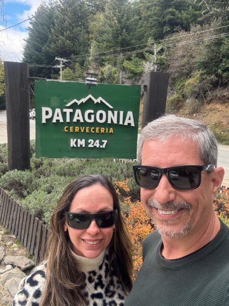 Uma mulher com blusa de estampa de leopardo e um homem sorridente de cabelos grisalhos posam para uma selfie em frente à placa verde da Patagonia Cerveceria, no quilômetro 24.7, cercados por árvores e vegetação.