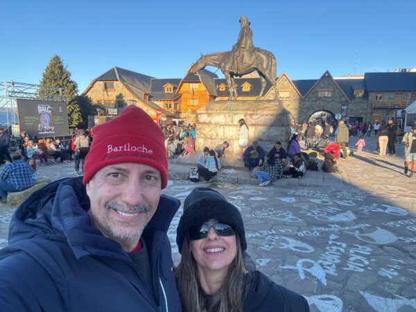 Selfie de um casal sorridente na praça do Centro Cívico de Bariloche, com o homem usando um gorro vermelho escrito Bariloche e a mulher um gorro preto, tendo ao fundo a estátua de bronze do General Roca a cavalo, edifícios históricos de pedra e madeira e várias pessoas sentadas na praça.