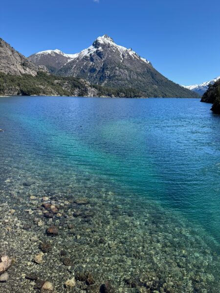 Vista panorâmica de um lago de águas extremamente transparentes e em tons de azul e turquesa, revelando pedras no fundo raso, cercado por montanhas arborizadas e um grande pico andino coberto de neve sob um céu azul limpo em Bariloche.