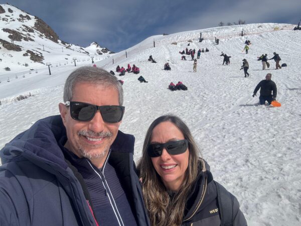 Selfie de um casal sorridente usando óculos de sol escuros e casacos de inverno pretos, com uma pista de esqui coberta de neve ao fundo, onde várias pessoas brincam e andam de esquibunda na montanha em Bariloche.