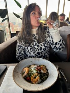 Clau Rousen vestindo blusa de estampa de leopardo enquanto bebe vinho branco em um restaurante sofisticado com vista para Santiago, Chile.