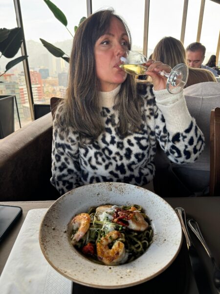 Clau Rousen vestindo blusa de estampa de leopardo enquanto bebe vinho branco em um restaurante sofisticado com vista para Santiago, Chile.