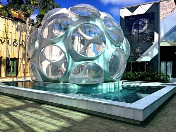 Uma grande cúpula esférica branca e futurista com janelas redondas transparentes (Fly's Eye Dome), instalada sobre um espelho d'água na praça Palm Court, no Miami Design District, cercada por fachadas de lojas de luxo e palmeiras sob um céu azul.