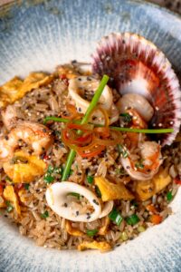 Arroz chaufa de frutos do mar servido com camarões, anéis de lula, vieira na concha, ovos mexidos, gergelim e cebolinha.