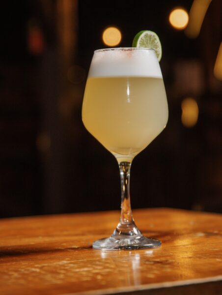 Coquetel Pisco Sour clássico servido em taça sobre mesa de madeira com espuma densa e rodela de limão na borda.