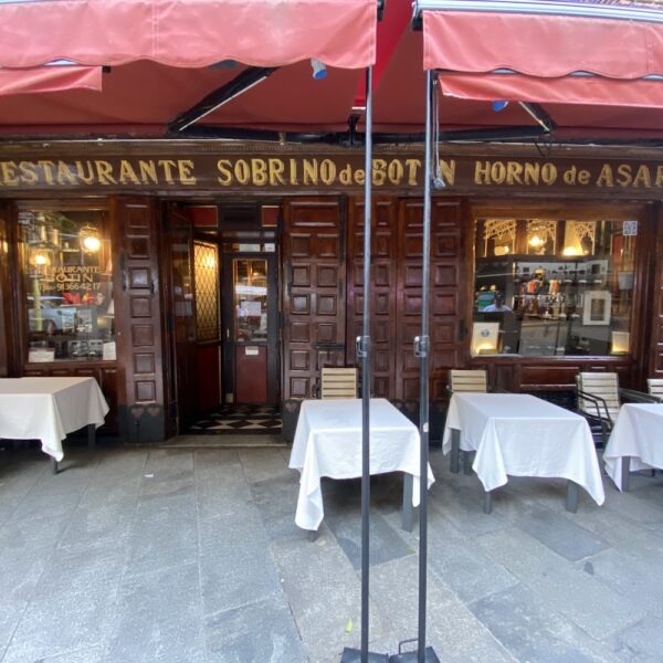 Fachada de madeira rústica do restaurante Sobrino de Botín em Madri, Espanha, exibindo o letreiro clássico dourado, vitrines antigas, toldos vermelhos e mesas com toalhas brancas na calçada.