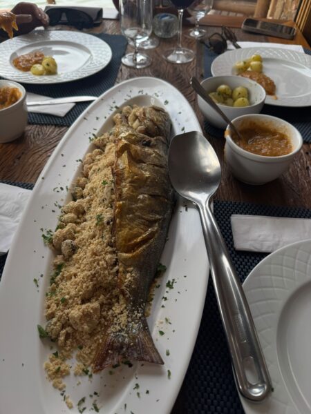 Anchova inteira assada servida em uma travessa branca, recheada com farofa de camarão, acompanhada de batatas cozidas e molho de camarão em potes sobre uma mesa de madeira.