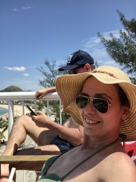 Mulher sorrindo de biquíni verde, óculos de sol e chapéu de palha, ao lado de um homem em uma sacada com vista para a praia.