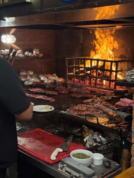 Parrilla tradicional do restaurante Fuego Celeste com chamas altas, diversos cortes de carne, linguiças e batatas assando na grelha.